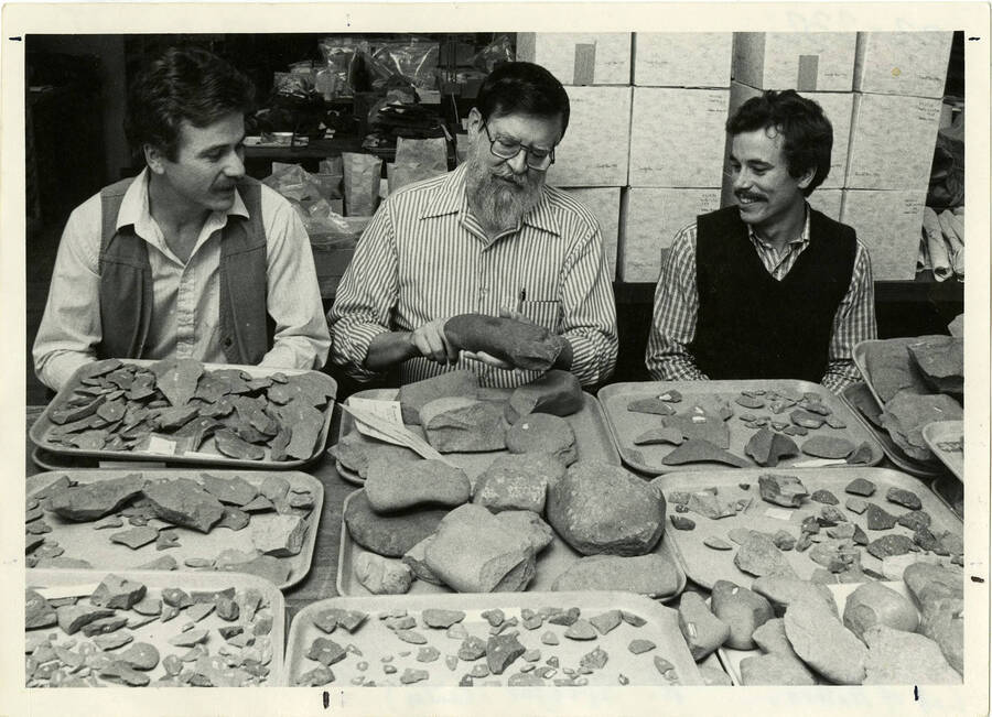 Three men pose for a photo in front of trays of rocks in the Lab of Anthropology. Dr. Roderick Sprague III is in the center.