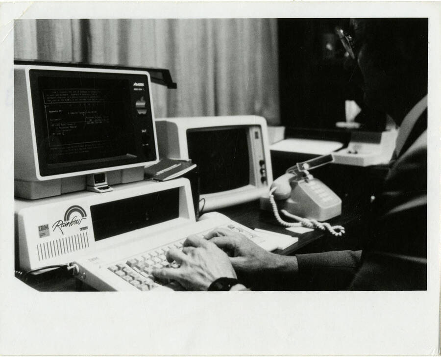 Man types on a computer on a desk. The computer monitor's label says ""Rainbow"" with ""IBM"" to the left of it.