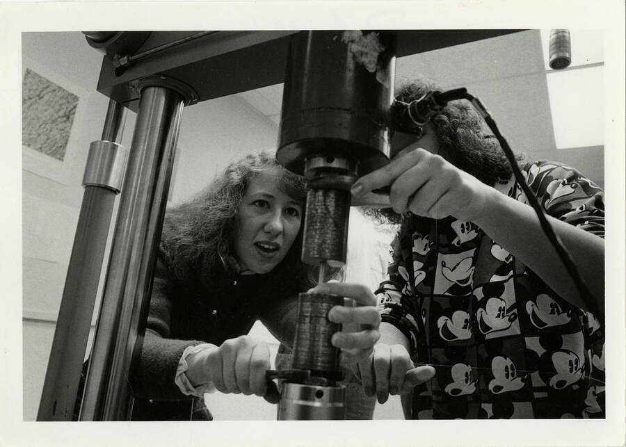 Two women using mechanical engineering equipment. Note on back of the photo says: ""Don't use! She should have protective glasses.""