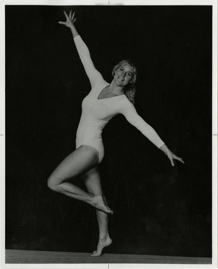 Woman posing in a long-sleeve white leotard and no shoes. Her body language, especially her pointed feet, depict her as a dancer.