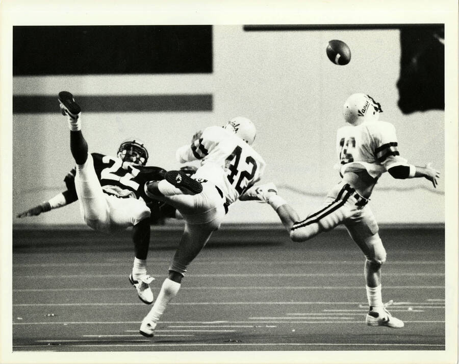 Idaho football player, wearing jersey number 42, helping player number 16 catch the football. Copy right note on back of photo reads: ""Copyright 1985 Paul C. Peck - This photograph cannot be copied, televised, reproduced, or used in any form without the express and written permission of Paul Peck Photography.""