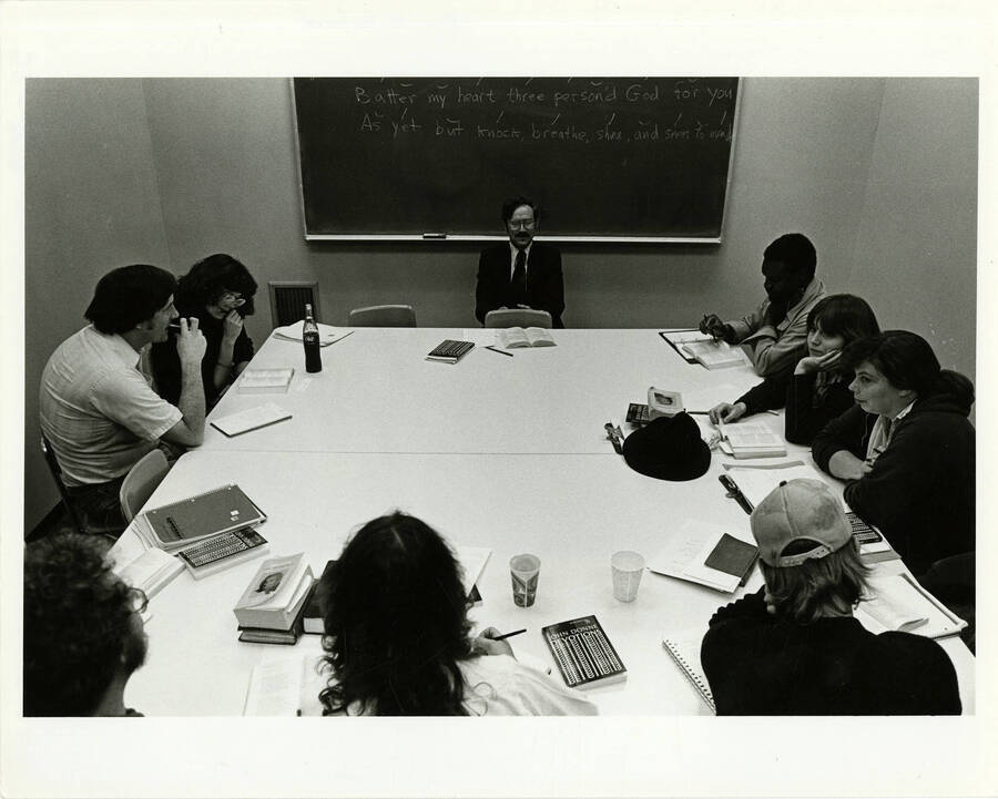 Ron McFarland teaching his poetry class. The chalkboard reads: ""Batter my heart three person'd God for you - As yet but knock, breathe, shine, and seek to mend."" Note the book John Donne Devotions and the Coke on the table. Note on the back of the photo reads: ""The University of Idaho is a modest size for a state university (more than 8,000 students), but an institution of vast range and resources in its programs and faculty -- from letters and science, education, business and economics, agriculture, art and architecture, mines and earth resources, engineering, forestry, and wildlife and range sciences, to law. The graduate school offers masters and doctoral degrees in many of these major areas. Leo Ames, Creative Director"". (Larger-sized duplicate of 52-251.)