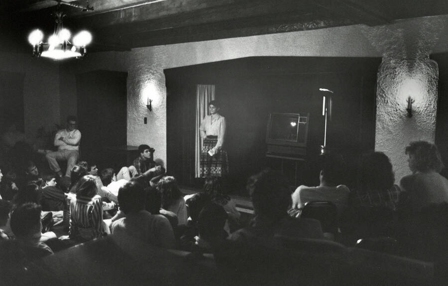 A woman standing in front of an audience, possibly performing or giving some kind of lecture. There is a large television in the background.