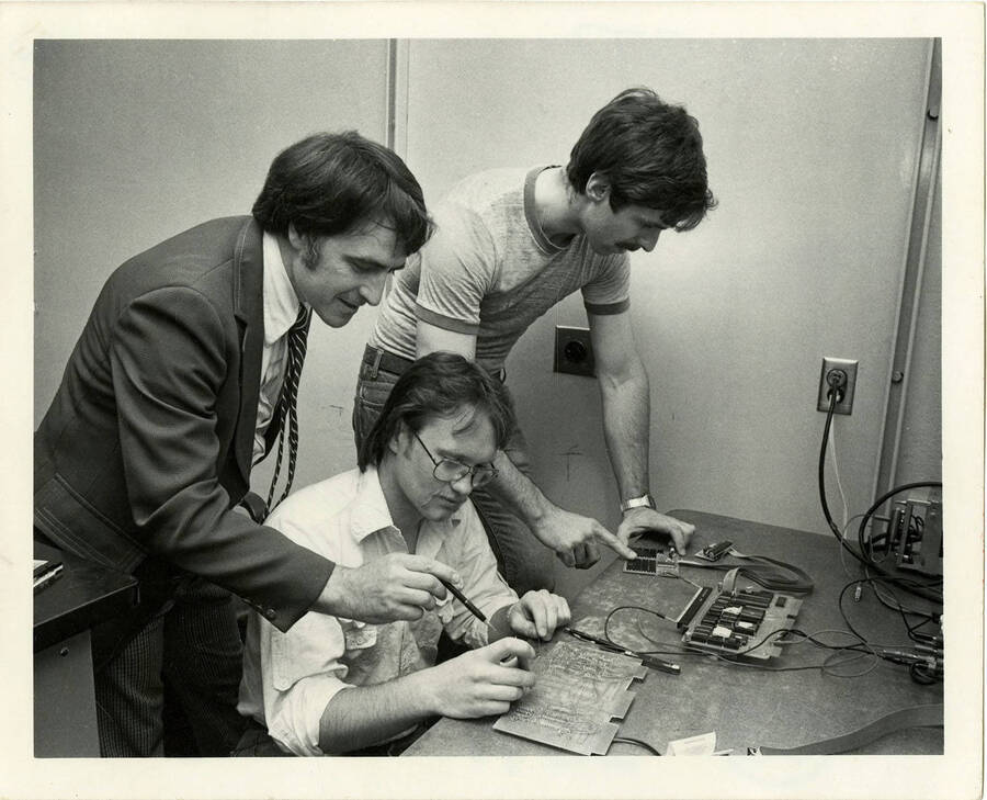 Three men working on electronic equipment on one desk.