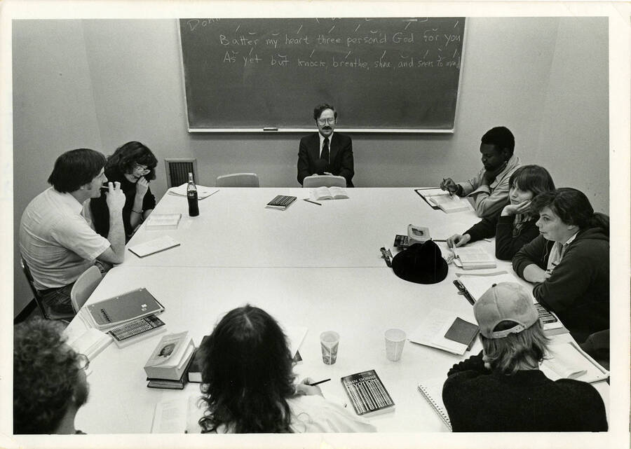 Ron McFarland teaching his poetry class. The chalkboard reads: ""Batter my heart three person'd God for you - As yet but knock, breathe, shine, and seek to mend."" Note the book John Donne Devotions and the Coke on the table.