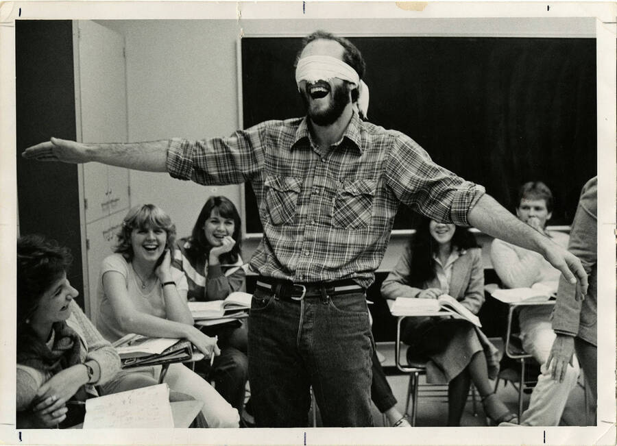 Blindfolded man standing in a classroom with students laughing at him.