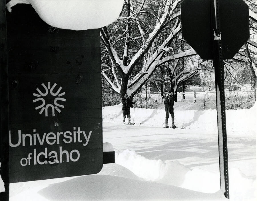 Two people cross-country skiing in the background of a shot of a U of I signage featuring the old ""UI star"" logo. A caption placard taped to the back of this photograph reads: ""University of Idaho, Publications Design - The University of Idaho is in northern Idaho which means winters will often be snowy. The University is in Moscow, a community of some 18,000 located at the eastern edge of the Palouse, wheat covered hills and near the foothills of the Bitterroot Mountains of the Rockies. Consequently, recreation opportunities vary widely. - Leo Ames, Creative Director""