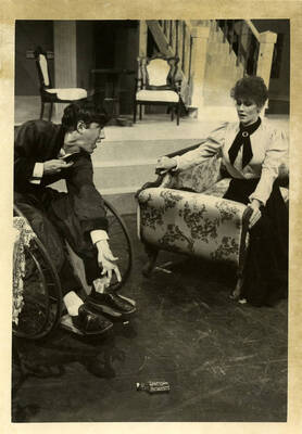 A man in a wheelchair reaching for a glass bottle while a woman sits on a couch and stares at it.