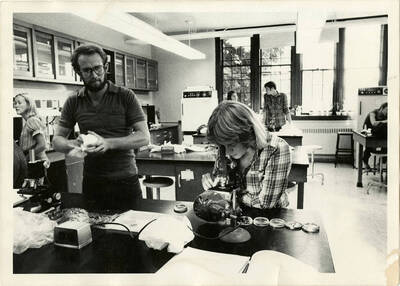 Biology lab students in the laboratory. One student examines petri dishes through a microscope. Another wipes a cup or bowl.