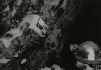 An individual using some kind of meter to record or observe data in a mine and wearing a hard hat with a head lamp.