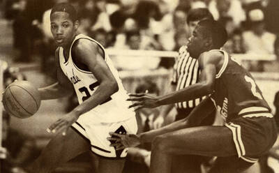 Two people cross-country skiing in the background of a shot of a U of I signage featuring the old ""UI star"" logo. A caption placard taped to the back of this photograph reads: ""University of Idaho, Publications Design - The University of Idaho is in northern Idaho which means winters will often be snowy. The University is in Moscow, a community of some 18,000 located at the eastern edge of the Palouse, wheat covered hills and near the foothills of the Bitterroot Mountains of the Rockies. Consequently, recreation opportunities vare widely. - Leo Ames, Creative Director""