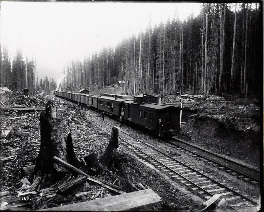 Image shows a Northern Pacific train stopped at the Coeur d'Alene Divide, on the Idaho-Montana border. Several railroad employees are posing on board and outside the train. A sign by the tracks says "Coeur d'Alene Divide Idaho/Montana elevation 4680 ft above sea level."