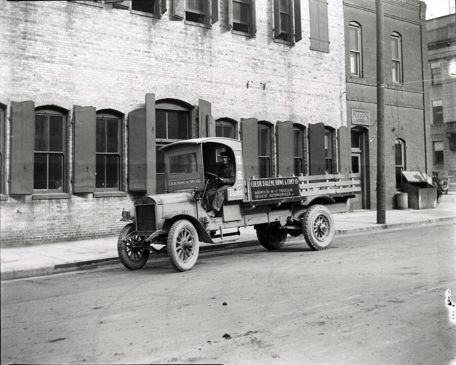 Image shows a man sitting in a GMC truck with advertising for the Coeur d'Alene Hardware and Foundry Company in Wallace, Idaho.