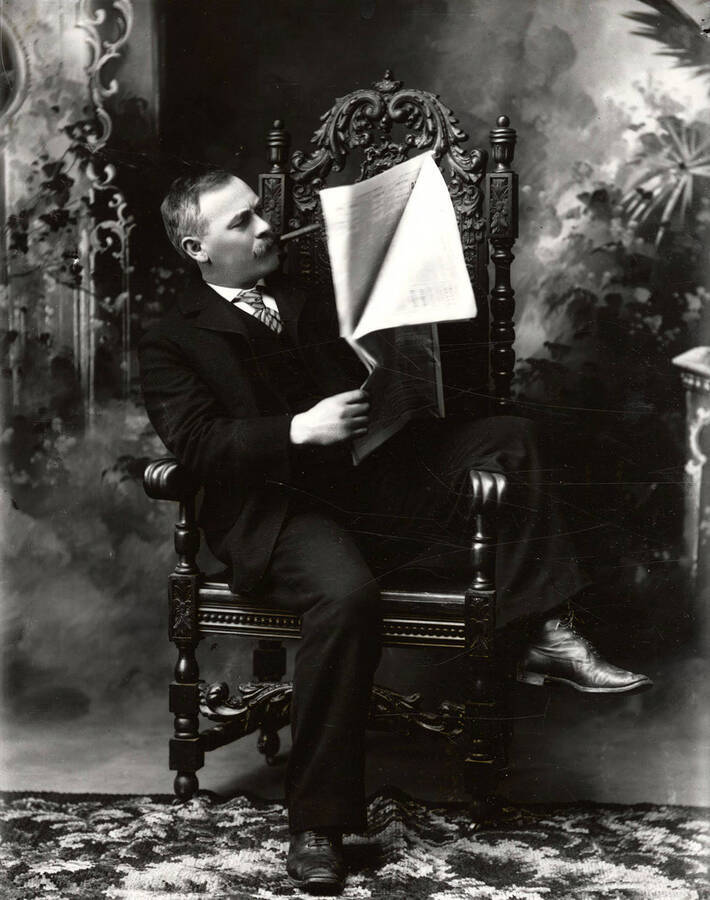 Portrait of Thomas Nathan Barnard (1861-1916) in a studio chair, reading newspaper, with a cigar in his mouth.