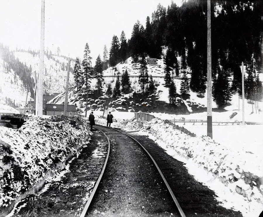 This image show two men posing next to O.W. R. & N. railroad tracks. Caption on front: "O.W.R. and N. Accident Case, 1913 Looking up Burke Canyon."