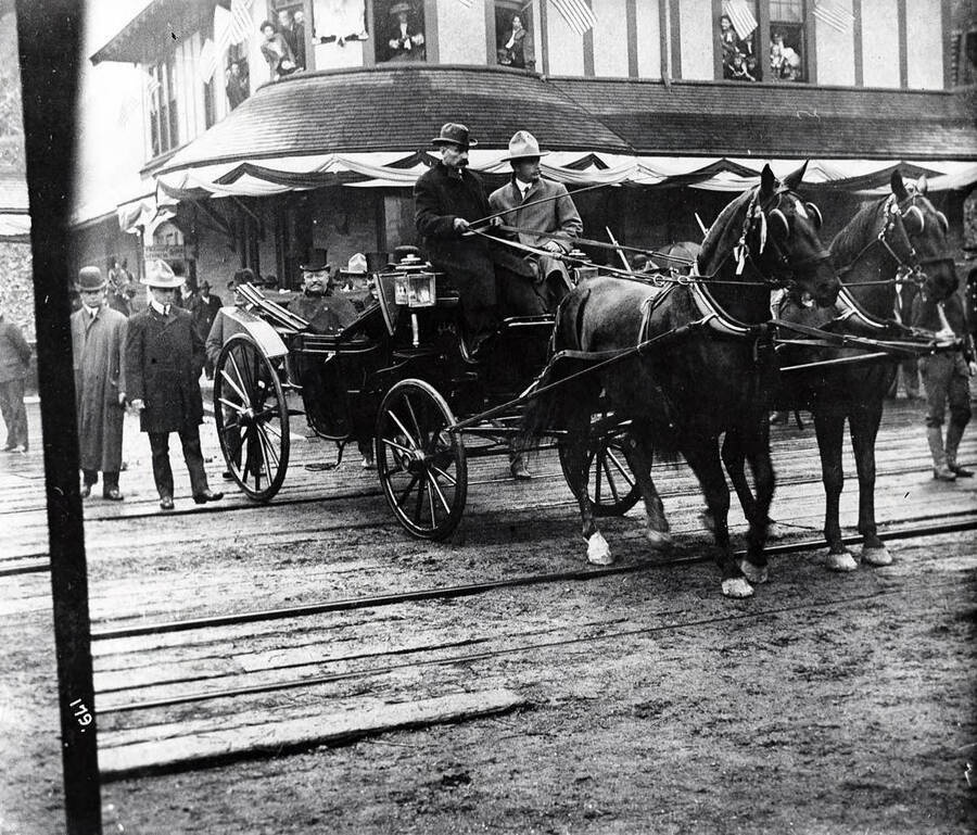 Image of President Theodore Roosevelt's visit to Wallace. He is shown sitting in the back of a horse-drawn carriage, surrounded by on-lookers on the street and in the windows of near-by buildings. Also shown in the carriage, are N.P. depot driver, Joe McDonald; Pat McGovern, chief of police and Harry W. McKinley.