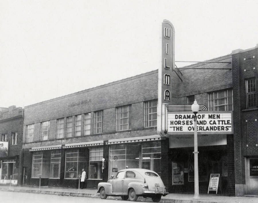 View of Wilma Theatre in Wallace, Idaho. A man is standing outside the theatre and a car is parked on the street. A real estate view taken for Idaho First National Bank.