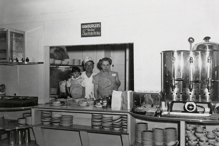 A man, woman, and child standing in front of the kitchen at the Silvio Cafe in Wallace, Idaho. A sign reading "Hamburgers 'to go' -- Buy 'em by the bag." hangs above them.