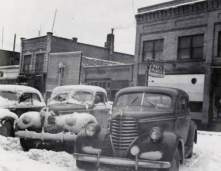 Snow-covered cars parked on Cedar Street in Wallace, Idaho. Pat's Place and other businesses can be seen behind the cars.