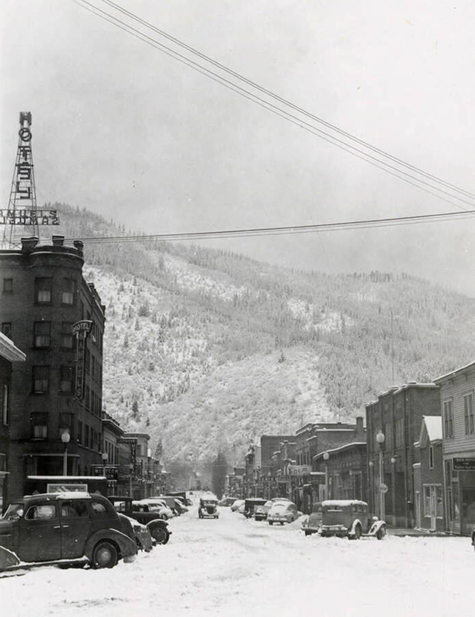 View of Cedar Street in Wallace, Idaho. Cars covered in snow are parked along the street, in front of buildings.