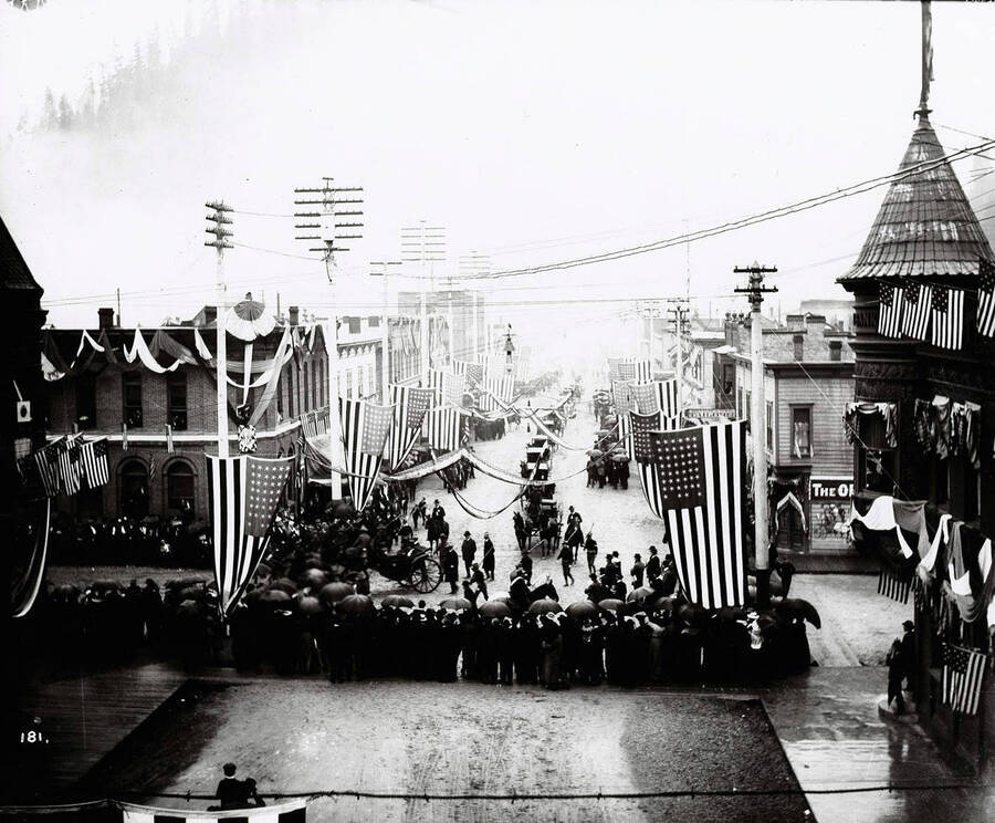 A panoramic view of the parade procession accompanying the visit of President Theodore Roosevelt. This image shows the corner of 6th and Bank Streets in Wallace, Idaho.