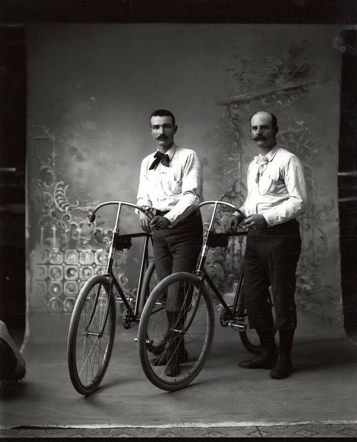Portrait of Mr. Ebley and Mr. Sherman, each standing beside a bicycle.