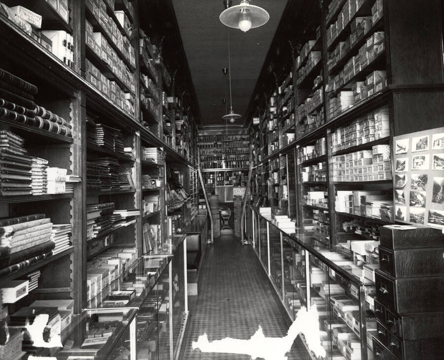 Interior view of the Wallace Drug Company in Wallace, Idaho. Along the sides, there are shelves reaching from floor to ceiling stocked with paper products and other items.