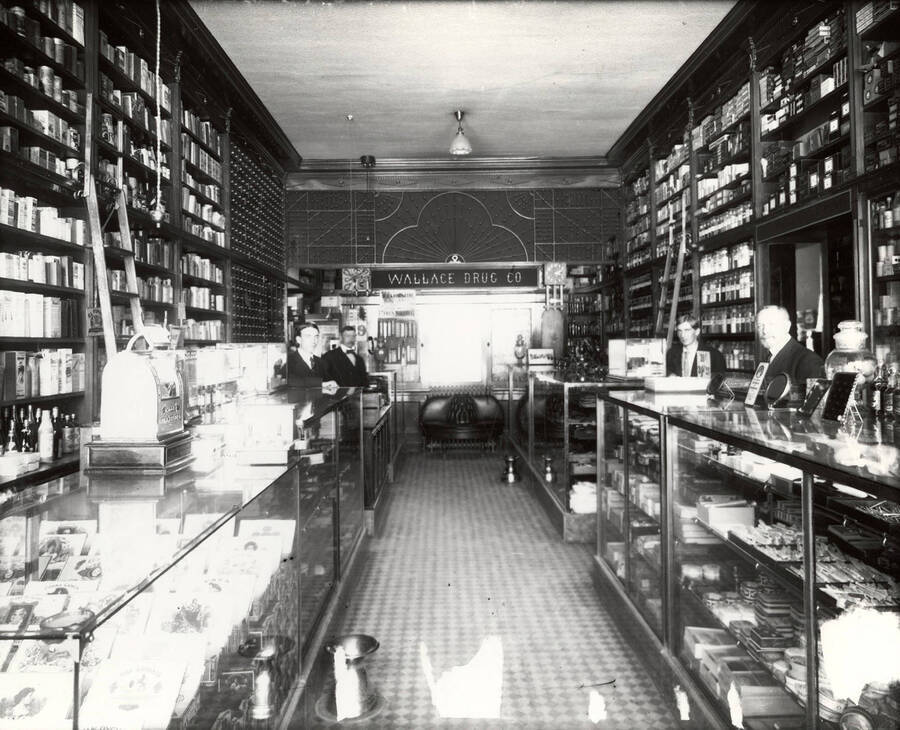 Interior view of the Wallace Drug Company in Wallace, Idaho. Along the sides, there are shelves reaching from floor to ceiling and four men standing behind display cases.