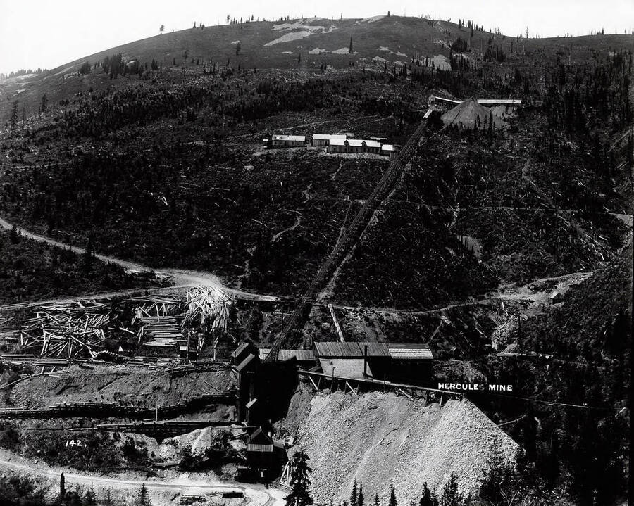 Hercules Mine in Burke, Idaho. Along with the surface structures, and waste dumps, logs are piled along the hillside, likely for use in the mining operations; Caption on front: "Hercules Mine."