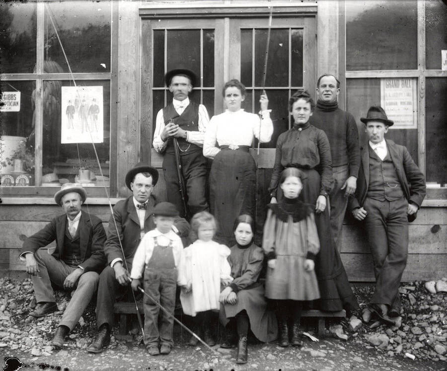 Five men, two women, and four children with fishing poles and a rifle sitting and standing in front of a store, possibly in Wallace, Idaho.