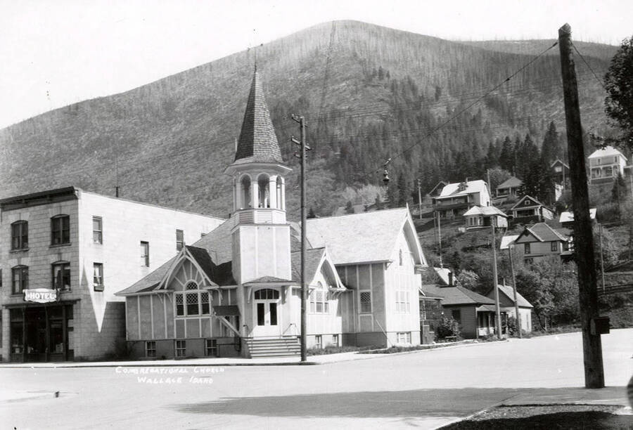 Exterior view of the Congregational Church in Wallace, Idaho. There is a hotel on its left and houses on the hill behind it.