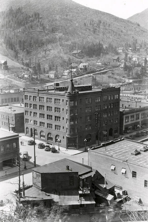 Exterior view of Samuels Hotel, with cars parked in front of it and other buildings and businesses surrounding it, in Wallace, Idaho. Barnard studio is the first window on the first floor to the right of the Samuel's Hotel