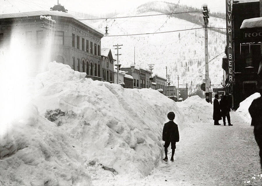 Looking down Cedar Street  during the winter in Wallace, Idaho. Residents stand around next to buildings in the snow.