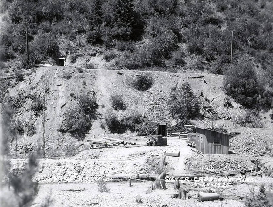 View of Silver Crescent Mine in Kellogg, Idaho. There are two men sitting by a pile of logs.