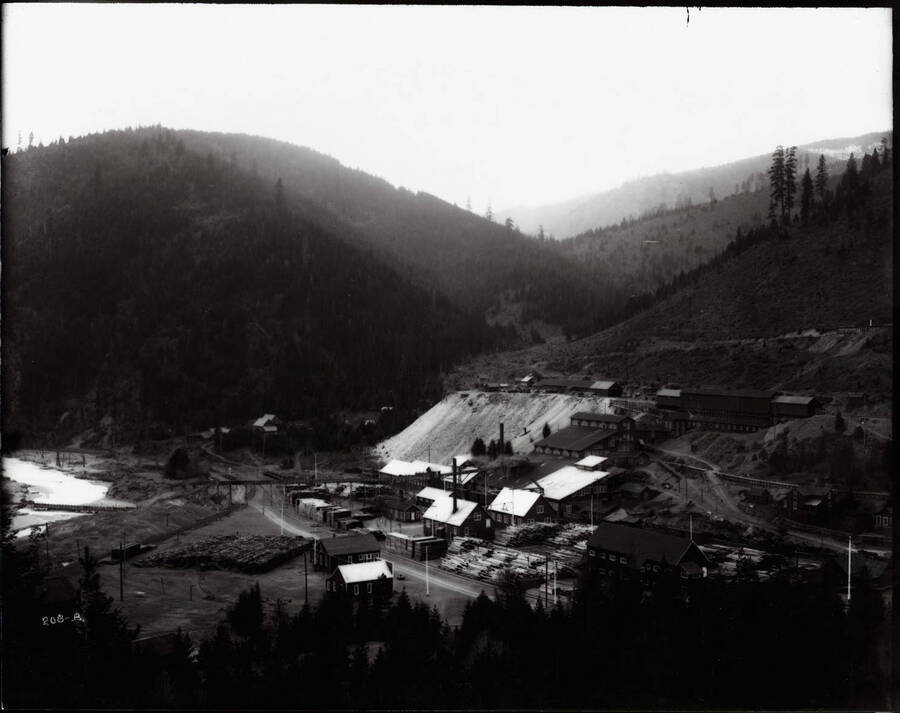 Image of the Morning Mill. Wooden buildings, mine waste dump, stacks of lumber and piles logs surround the area.