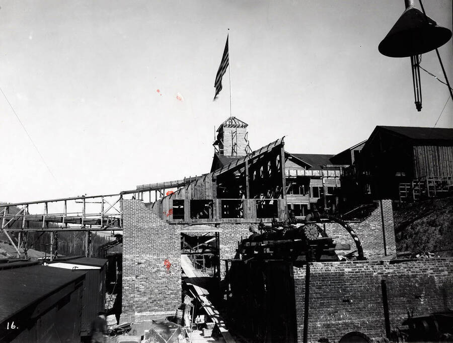 Image shows brick buildings and rail cars at the Bunker Hill and Sullivan Mill in Wardner, Idaho; A United States flag is flying over one of the buildings.