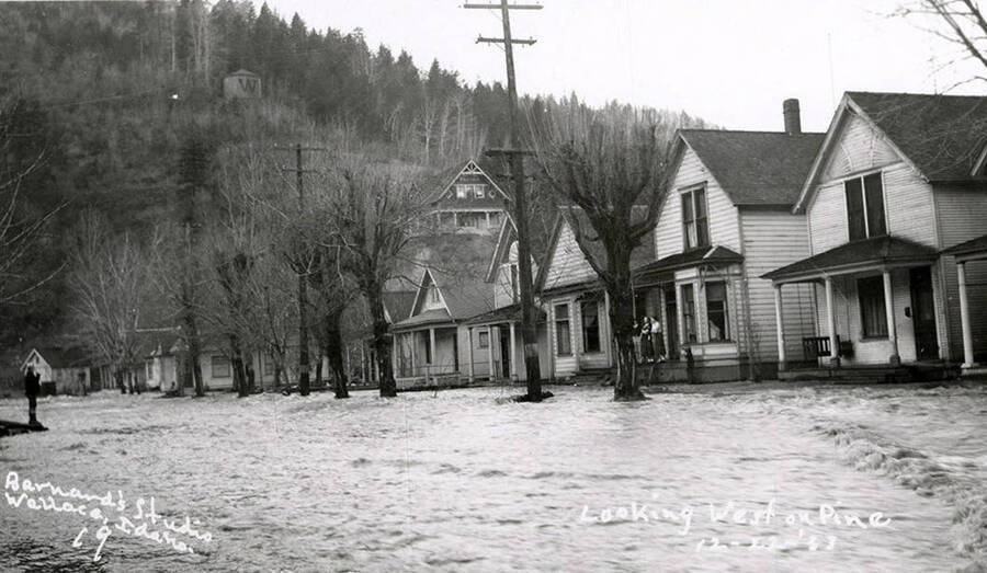 Looking west on Pine Street during the Placer Creek flood in Wallace, Idaho. Houses line the street and some residents can be seen standing on porches.