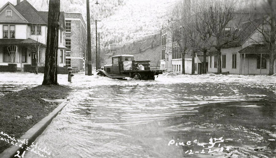 View of Pine and Third Street during the Placer Creek flood in Wallace, Idaho. Houses line the street and a truck sits in the middle of the street.