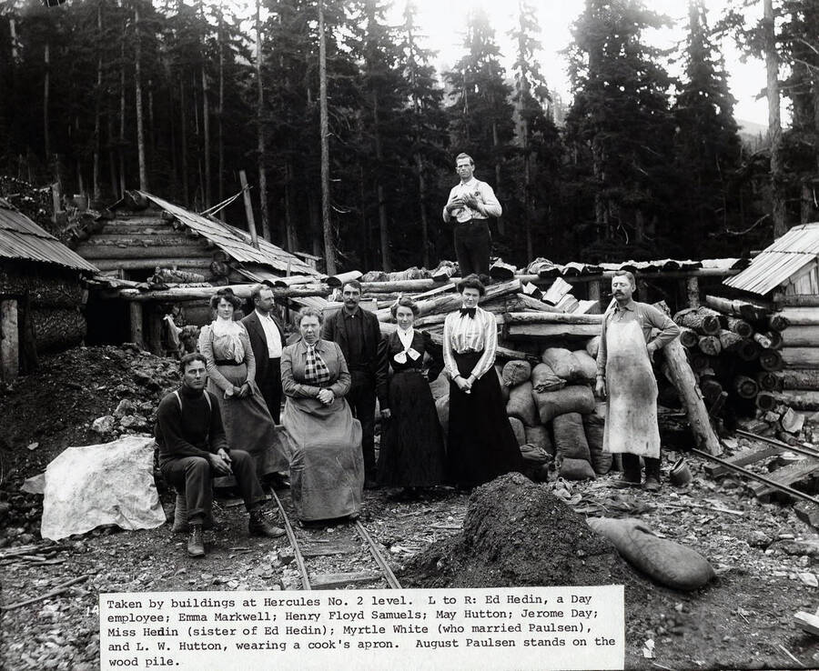 Group of nine men and women in front of the Hercules Mine, shortly after its discovery; Caption on Front: Taken by buildings at Hercules No. 2 level. L to R: Ed Hedin, a Day employee; Emma Markwell; Henry Floyd Samuels; May Hutton; Jerome Day; Miss Hedin (sister of Ed Hedin); Myrtle White (who married Paulsen), and L. W. Hutton, wearing a cooks apron. August Paulsen stands on the wood pile.