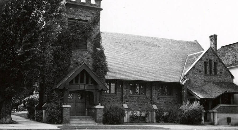 Exterior view of the Holy Trinity Episcopal Church in Wallace, Idaho. There are vines running up the side of the building and a large tree on the left.