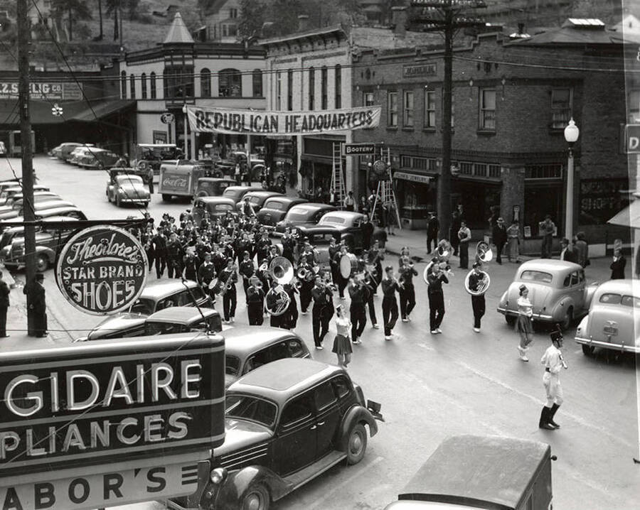 The Wallace High School Band marching in a parade in Wallace, Idaho. Cars are parked in front of buildings and stores that line the street.