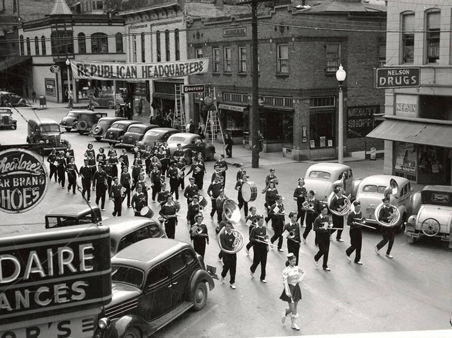 The Wallace High School Band marching in a parade in Wallace, Idaho. Cars are parked in front of buildings and stores that line the street.