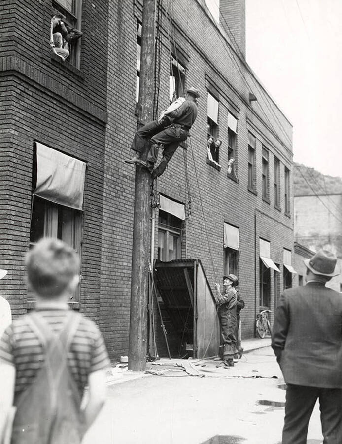 Showing a rescue demonstration during the Fire Chief's Convention in Wallace, Idaho. A man bringing a person down a ladder on the side of the building, while people watch from windows and from the ground.