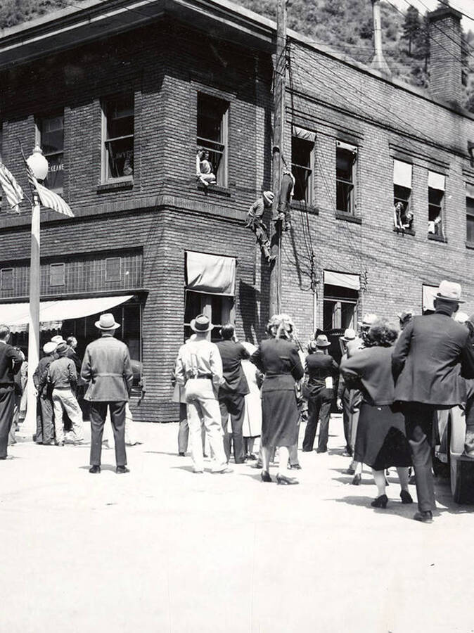 Showing a rescue demonstration during the Fire Chief's Convention in Wallace, Idaho. Two men climb a pole on the side of a building, while people watch from windows and from the ground.