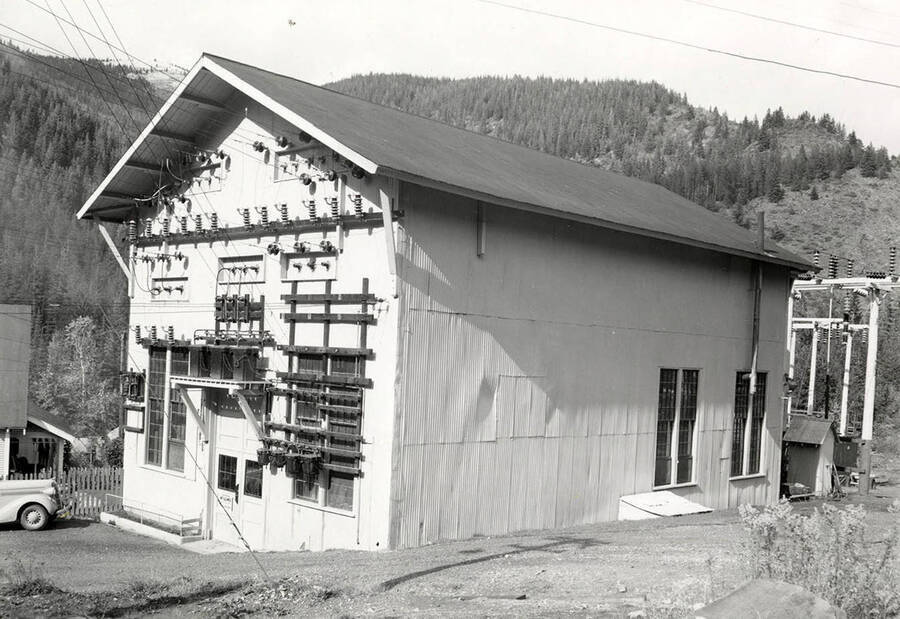 Exterior view of the Montana Power Company substation in Wallace, Idaho. Hills can be seen in the background and wires are running to and from the building.
