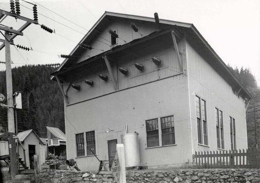 Exterior view of the Montana Power Company substation in Wallace, Idaho. Hills can be seen in the background and wires are running to and from the building.
