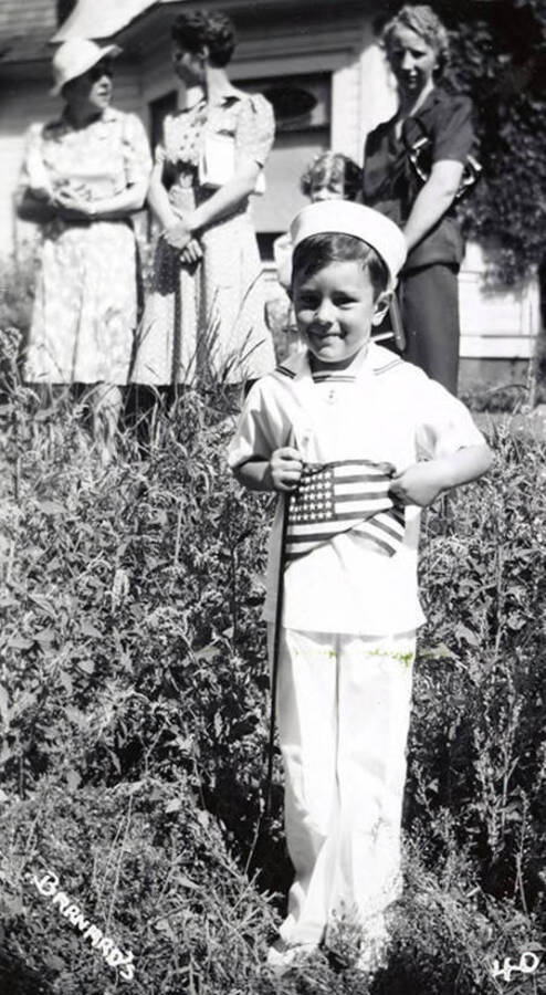A boy, holding a small flag, in costume for the Mullan 49'er parade in Mullan, Idaho. Three women stand behind him.