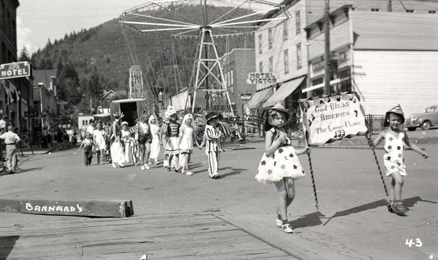Children, who are in costume, walking in the Mullan 49'er parade in Mullan, Idaho. Two girls walk in front, holding a sign that says "God Bless America The Land I Love."
