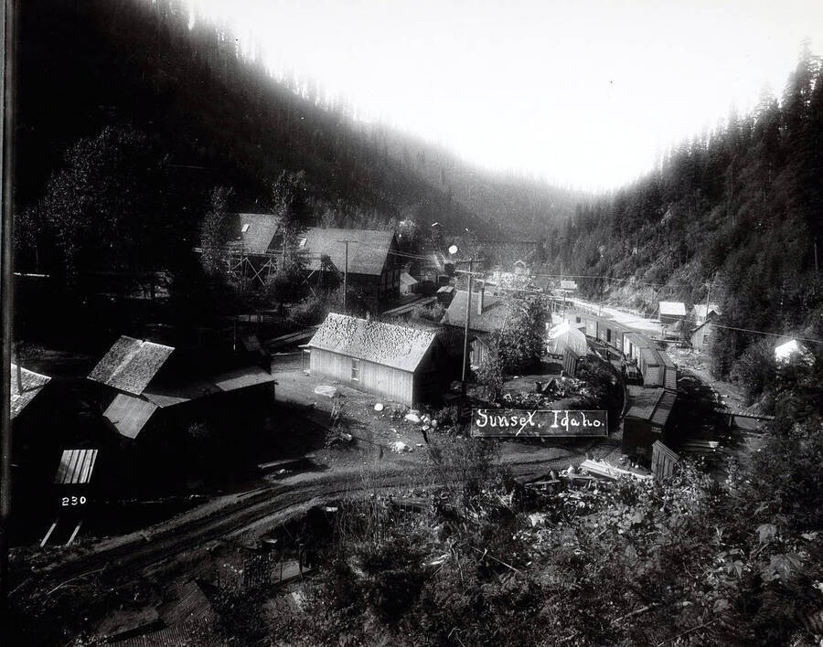 Caption on front: "Sunset, Idaho." Image shows a variety of buildings. Railroad boxcars line the tracks. A wooden railroad trestle can be seen in the distance.