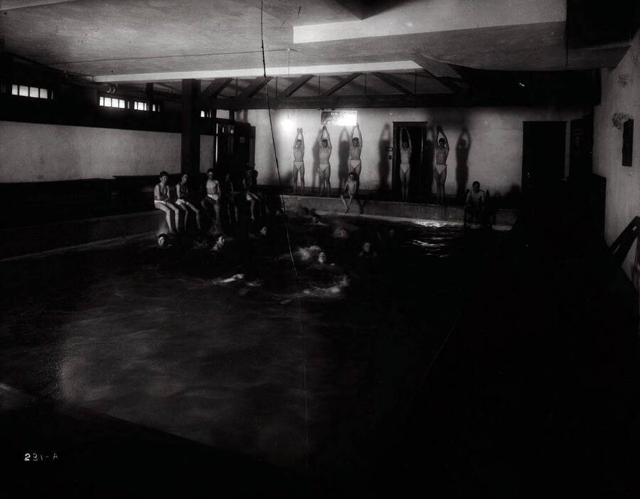 Interior image of the Wallace High School swimming pool. This photo shows several male students posing in diving positions, sitting around and swimming in the pool.